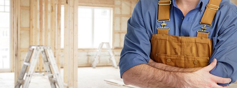 man wearing bib overalls standing with crossed arms in home under construction